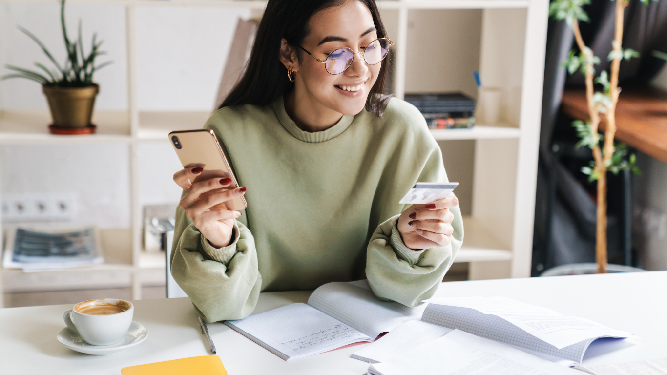 Mulher sorrindo sentada em uma mesa com cartão de crédito em uma mão e celular em outra.